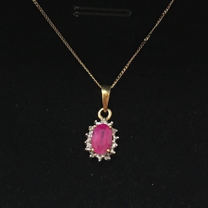 May include: A gold necklace with a pendant featuring a large, oval-shaped ruby surrounded by small, clear gemstones. The ruby is a vibrant pink color, and the necklace is set against a black background.