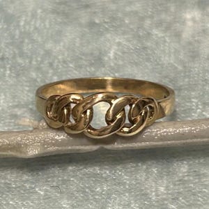 May include: A gold-colored ring with a chain link design. The ring features three interconnected chain links across the top, creating a bold and stylish look. The ring is set against a light blue background.