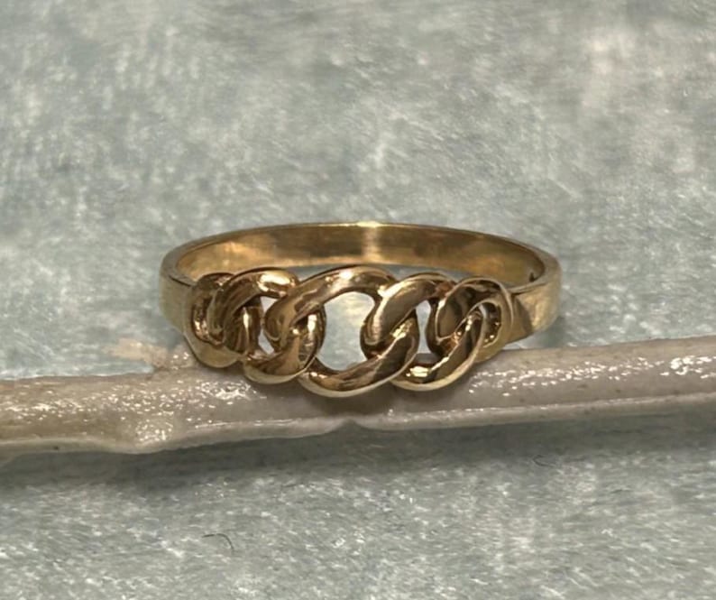 May include: A gold-colored ring with a chain link design. The ring features three interconnected chain links across the top, creating a bold and stylish look. The ring is set against a light blue background.