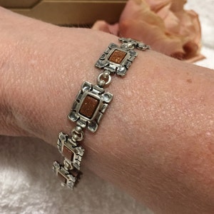 May include: A silver bracelet with square-shaped brown sunstone accents. The bracelet is made of linked silver squares with a textured surface.