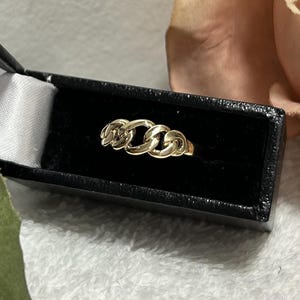 May include: A gold chain link ring displayed in a black jewelry box with a white satin interior. The ring features three interconnected chain links. The background includes a soft pink flower and a green fabric.