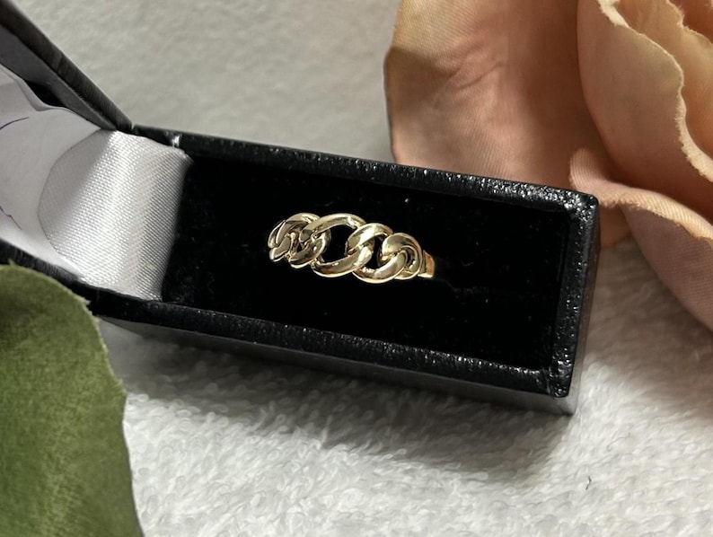 May include: A gold chain link ring displayed in a black jewelry box with a white satin interior. The ring features three interconnected chain links. The background includes a soft pink flower and a green fabric.