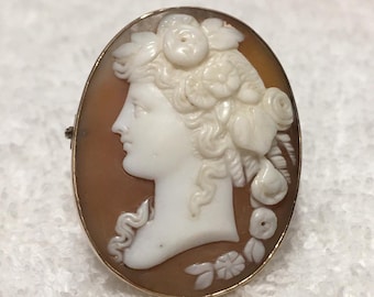 May include: Oval cameo brooch featuring a profile of a woman with floral accents in white relief against a brown background. The cameo is set in a gold-colored frame, showcasing a classic and elegant design.