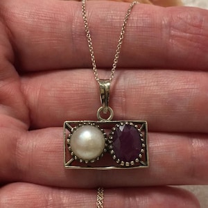 Op de afbeelding: Een zilveren ketting met een rechthoekige hanger met een parel en een robijn. De parel is wit en rond, de robijn is dieprood en facetgeslepen. De ketting is delicaat.