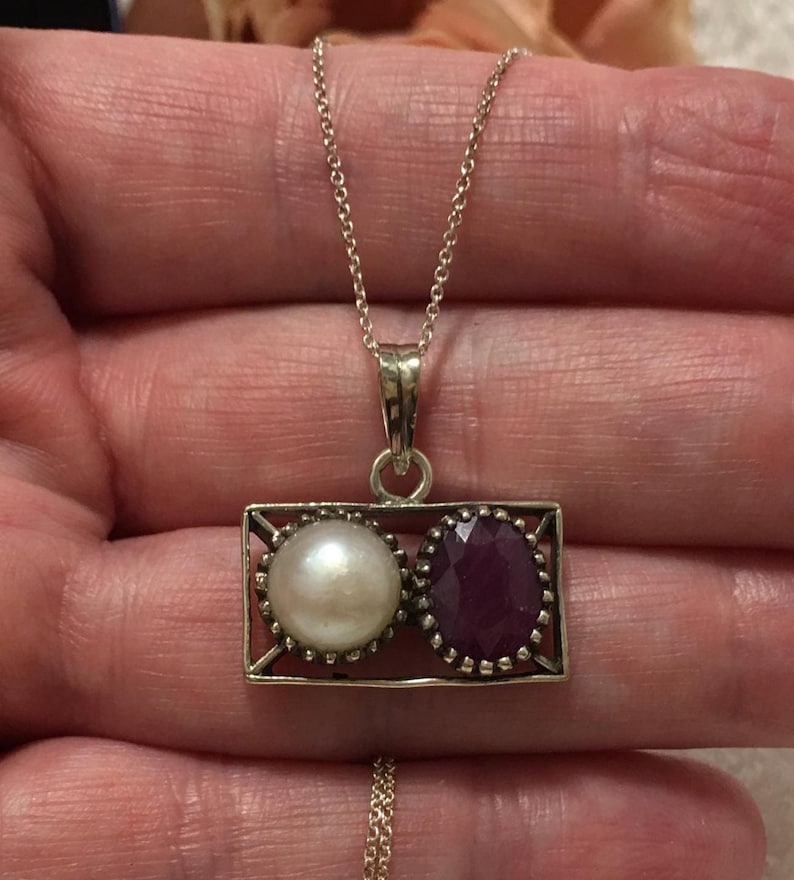 Op de afbeelding: Een zilveren ketting met een rechthoekige hanger met een parel en een robijn. De parel is wit en rond, de robijn is dieprood en facetgeslepen. De ketting is delicaat.