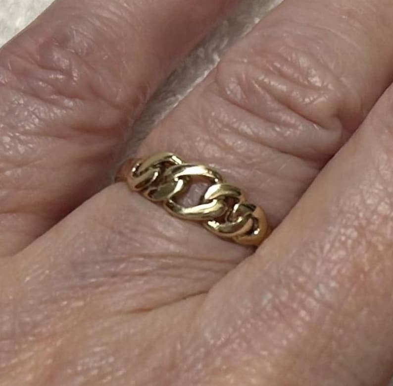 May include: A gold-colored ring with a chain link design. The ring is made of a shiny, reflective metal and is worn on a finger. The ring's design is a series of interconnected links.