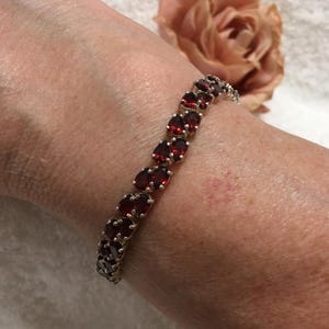 Könnte beinhalten: Ein silbernes Armband mit ovalen, tiefroten Edelsteinen. Das Armband wird an einem Handgelenk getragen, mit einer verschwommenen Rose im Hintergrund. Die Edelsteine sind in einem silbernen Band gefasst, was einen klassischen und eleganten Look erzeugt. Das Armband ist ein Schmuckstück.