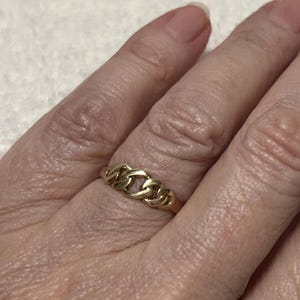May include: A gold-colored ring with a chain link design. The ring is worn on a finger and appears to be made of metal. The ring's links are interlocked, creating a continuous pattern. The ring is a piece of jewelry.