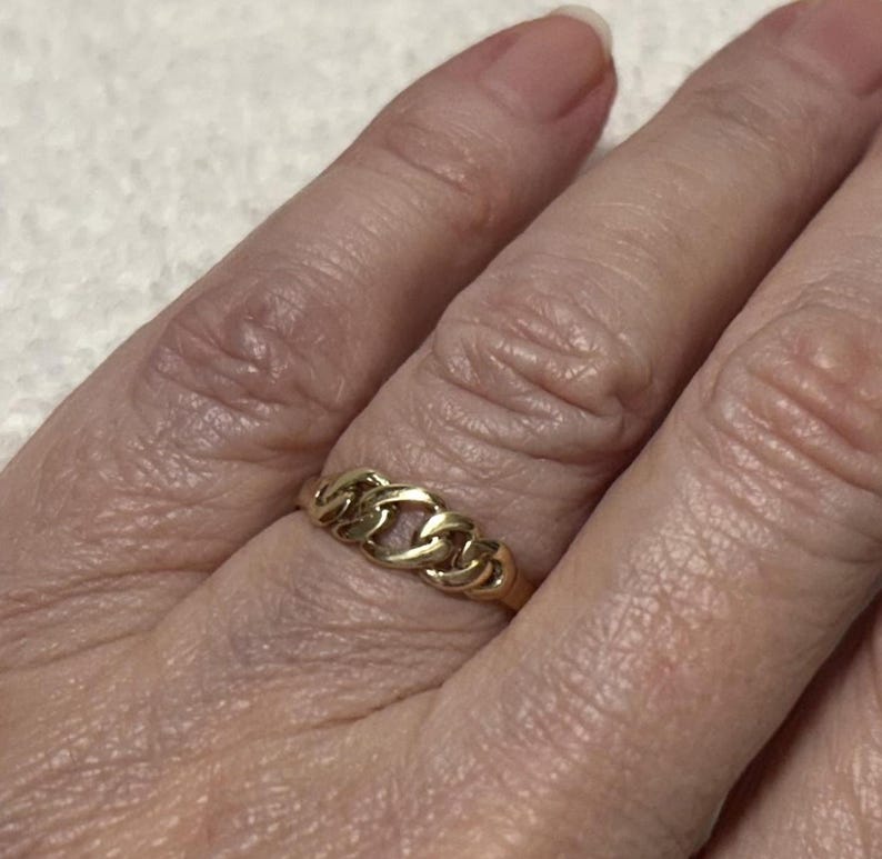 May include: A gold-colored ring with a chain link design. The ring is worn on a finger and appears to be made of metal. The ring's links are interlocked, creating a continuous pattern. The ring is a piece of jewelry.