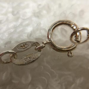 Op de afbeelding: Close-up van een zilveren ketting sluiting en ketting. De sluiting is een cirkelvormig veeringtype, verbonden met een klein ovaal label met de stempel "925 ITALY". De ketting is delicaat en de achtergrond is een zachte, witte, fuzzy textuur.