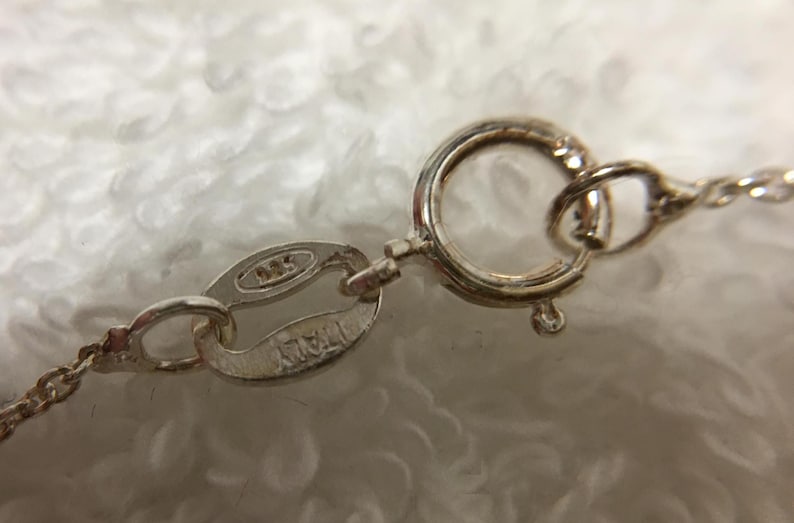 Op de afbeelding: Close-up van een zilveren ketting sluiting en ketting. De sluiting is een cirkelvormig veeringtype, verbonden met een klein ovaal label met de stempel "925 ITALY". De ketting is delicaat en de achtergrond is een zachte, witte, fuzzy textuur.