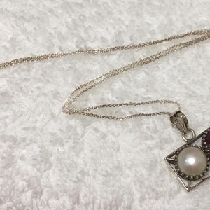 Op de afbeelding: Zilveren ketting met een rechthoekige hanger met een witte parel en een dieprode edelsteen. De hanger is omlijst door een zilveren rand en hangt aan een fijne zilveren ketting.