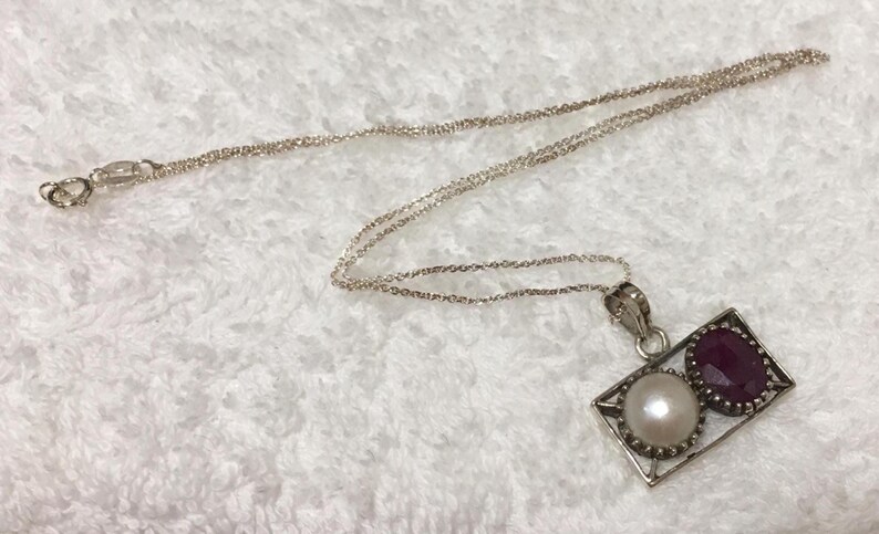 Op de afbeelding: Zilveren ketting met een rechthoekige hanger met een witte parel en een dieprode edelsteen. De hanger is omlijst door een zilveren rand en hangt aan een fijne zilveren ketting.