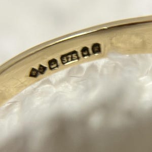 May include: Close-up of a gold ring with hallmarks. The ring's band is smooth and shiny, with several small stamps including the number "375". The ring is set against a soft, white background.