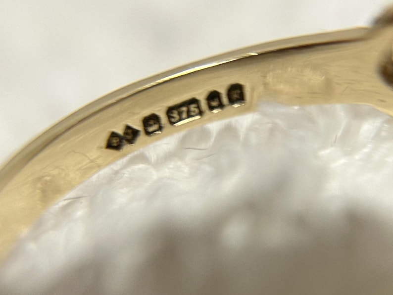 May include: Close-up of a gold ring with hallmarks. The ring's band is smooth and shiny, with several small stamps including the number "375". The ring is set against a soft, white background.