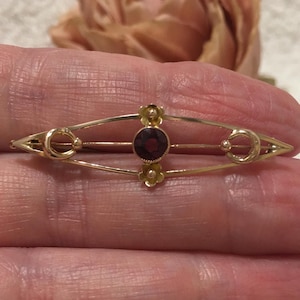 Puede incluir: Broche dorado con una gema central redonda de color rojo oscuro. El broche tiene un diseño simétrico con detalles florales y elementos curvos. El broche está sostenido en una mano.