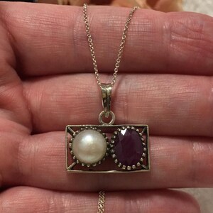 Op de afbeelding: Zilveren ketting met een rechthoekige hanger met een witte parel en een gefacetteerde, dieprode edelsteen. De hanger hangt aan een fijne ketting. De parel is rond en glanzend, de edelsteen is briljant geslepen.