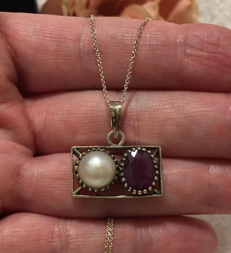 Op de afbeelding: Zilveren ketting met een rechthoekige hanger met een witte parel en een gefacetteerde, dieprode edelsteen. De hanger hangt aan een fijne ketting. De parel is rond en glanzend, de edelsteen is briljant geslepen.