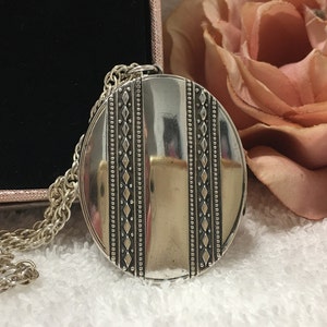 May include: A silver oval locket with a chain. The locket has a decorative border with a repeating pattern of small circles and diamonds. The locket is closed and has a smooth, reflective surface.