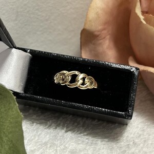 May include: A gold chain link ring displayed in a black jewelry box with a white satin lining. The ring features three interconnected chain links. The background includes a soft pink flower and a white, textured surface.