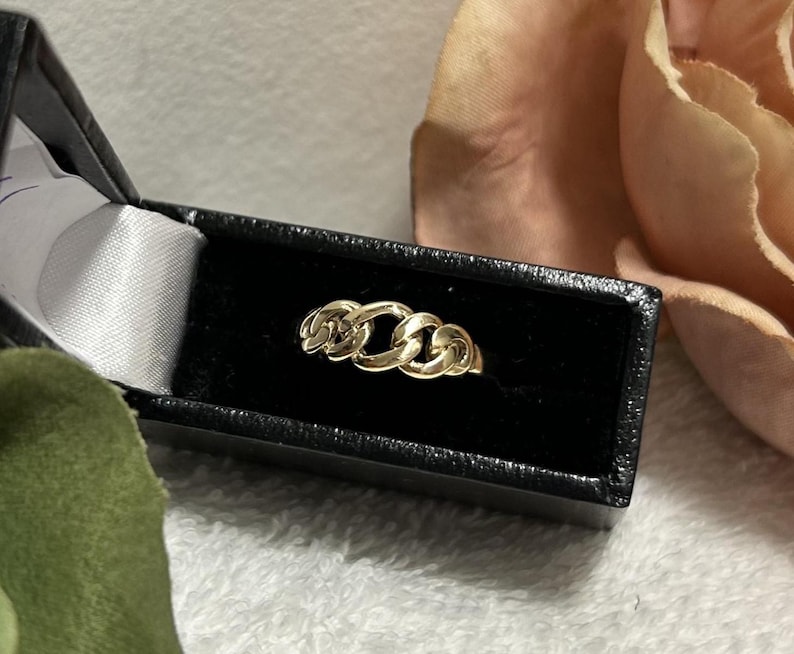 May include: A gold chain link ring displayed in a black jewelry box with a white satin lining. The ring features three interconnected chain links. The background includes a soft pink flower and a white, textured surface.
