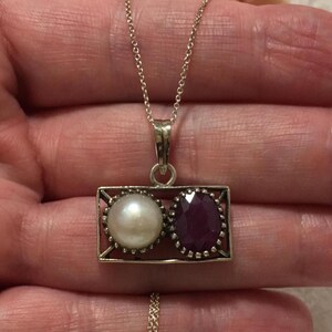 Op de afbeelding: Zilveren hangerketting met een rechthoekig frame met een parel en een robijn. De parel is rond en wit, de robijn ovaal en dieprood. De ketting heeft een fijne zilveren ketting.