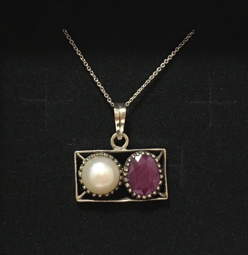 Op de afbeelding: Zilveren hangerketting met een parel en een robijn. De hanger heeft een rechthoekig frame, met een parel links en een robijn rechts. De ketting is delicaat en zilverkleurig.