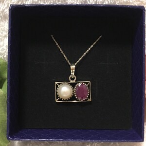 Op de afbeelding: Een zilveren ketting met een rechthoekige hanger met een witte parel en een rode edelsteen. De ketting wordt gepresenteerd in een donkerblauwe doos met een zwart interieur, tegen een zachte, getextureerde achtergrond.