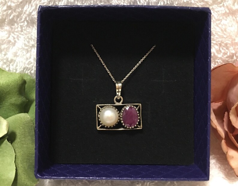 Op de afbeelding: Een zilveren ketting met een rechthoekige hanger met een witte parel en een rode edelsteen. De ketting wordt gepresenteerd in een donkerblauwe doos met een zwart interieur, tegen een zachte, getextureerde achtergrond.