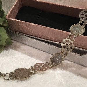 May include: A silver bracelet with a chain and coin-shaped charms. The charms have a floral design and a profile of a person on them.
