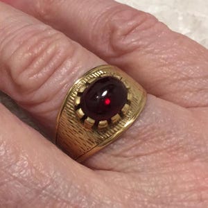Op de afbeelding: Een goudkleurige ring met een getextureerde band en een grote, ovale, donkerrode edelsteen. De edelsteen is gezet in een decoratieve bezel. De ring wordt aan een vinger gedragen.