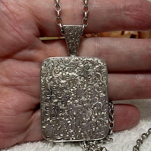 Könnte beinhalten: Silberfarbene rechteckige Anhänger-Halskette mit einem verzierten, wirbelnden Muster. Der Anhänger hängt an einer passenden Kette. Das filigrane Design bedeckt die gesamte Oberfläche des Anhängers und erzeugt eine Vintage-Ästhetik. Die Kette ist ca. 45 cm lang.