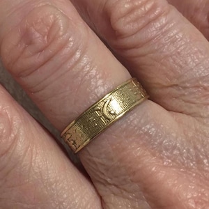 May include: A gold ring with a detailed pattern featuring crescent moon and star designs. The ring is worn on a finger, showcasing its intricate texture and design. The ring's band has a textured surface.