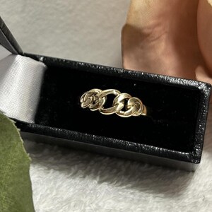 May include: A gold-colored ring with a chain link design, displayed in a black jewelry box with a white satin lining. The ring is the focal point, set against a soft, blurred background.