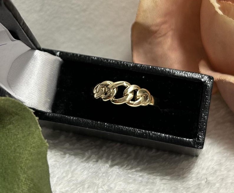 May include: A gold-colored ring with a chain link design, displayed in a black jewelry box with a white satin lining. The ring is the focal point, set against a soft, blurred background.