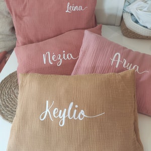 K&ouml;nnte beinhalten: Vier weiche, rosa und braune Kissen mit wei&szlig;en bestickten Namen: Leina, N&egrave;zia, Arna und Keylio. Die Kissen sind &uuml;bereinander gestapelt, wobei das braune Kissen unten liegt.