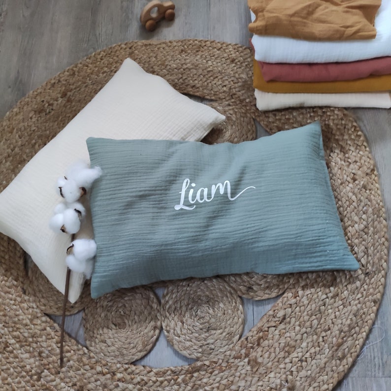 K&ouml;nnte beinhalten: Ein hellblaues Kissen mit dem Namen "Liam", der in Wei&szlig; auf einem nat&uuml;rlichen Jute-Teppich gestickt ist.