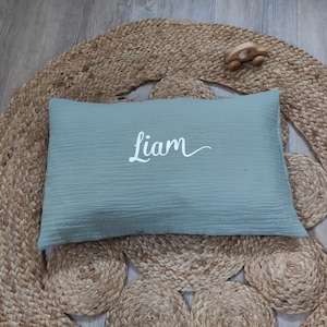 K&ouml;nnte beinhalten: Ein hellgr&uuml;nes Musselin-Kissen mit dem Namen "Liam", der in Wei&szlig; auf die Vorderseite gestickt ist. Das Kissen liegt auf einem gewebten Teppich.