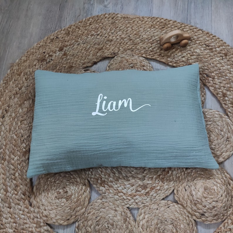 K&ouml;nnte beinhalten: Ein hellgr&uuml;nes Musselin-Kissen mit dem Namen "Liam", der in Wei&szlig; auf die Vorderseite gestickt ist. Das Kissen liegt auf einem gewebten Teppich.