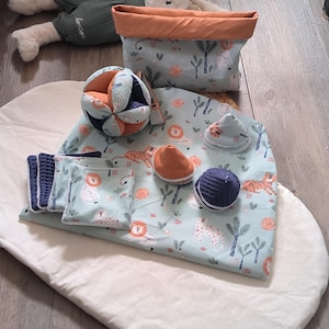 Op de afbeelding: Een verzameling babyartikelen, waaronder een speelmat, een stoffen mand en verschillende kleine speeltjes. De speelmat en mand hebben een lichtblauwe stof met een jungle-thema. De speeltjes omvatten een textuurbal en een zachte, gesegmenteerde bal.