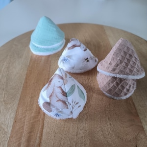 Puede incluir: Cuatro almohadillas de desmaquillaje reutilizables de tela en forma de cono. Una almohadilla es de color verde claro, otra es blanca con un patrón floral, otra es blanca con un patrón de conejo y la última es marrón con una textura de gofre.