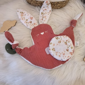 Peut inclure: Une doudou pour bébé en forme de lapin, de couleur corail, avec des motifs floraux sur les oreilles et un patch rond. Bordure blanche et détail cœur. Un nuage en bois est également visible.