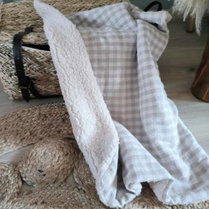 Peut inclure: Un panier en osier avec une sangle en cuir est posé à côté d'une couverture à carreaux beige et blanche et d'une couverture moelleuse crème. Les articles sont sur un tapis tissé, créant une scène aux tons neutres et chaleureux.