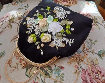 May include: A black velvet handbag featuring a gold-toned metal frame and a delicate chain strap. The front is decorated with intricate ribbon embroidery in shades of white, cream, and sage green, accented with small white pearls and clear crystals.