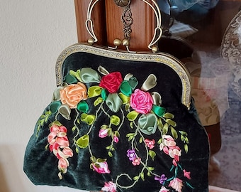 May include: A dark green velvet handbag with a brass frame and a floral design embroidered with silk ribbon. The flowers are pink, red, and peach.