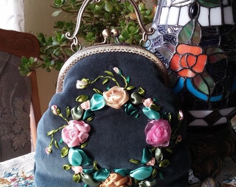 May include: A teal velvet handbag with a floral ribbon design. The bag features a decorative metal handle and clasp. The ribbon flowers are pink, peach, and gold, with green and teal ribbon leaves. The bag is approximately 8 inches wide.