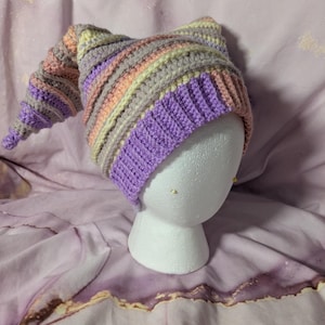 May include: A handmade crochet hat in a whimsical style, featuring a long, pointed tip. The hat showcases a striped pattern in shades of purple, pink, gray, and yellow. The hat is displayed on a white mannequin head.