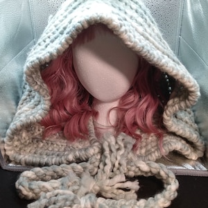 May include: A hooded cowl made of chunky yarn in a light blue and white color combination. The cowl is shown on a mannequin with pink hair. The cowl has a long, loose scarf attached.