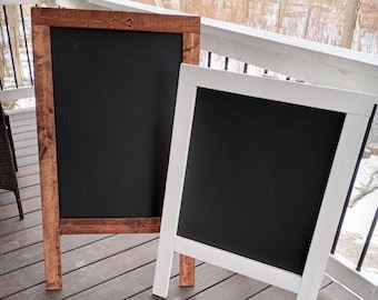 May include: Two wooden A-frame chalkboard signs. One sign is stained a dark brown and the other is painted white. Both signs have black chalkboard inserts.