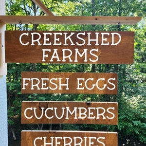 May include: A wooden sign with four hanging panels. The top panel reads "Creekshed Farms". The other panels read "Fresh Eggs", "Cucumbers", and "Cherries".